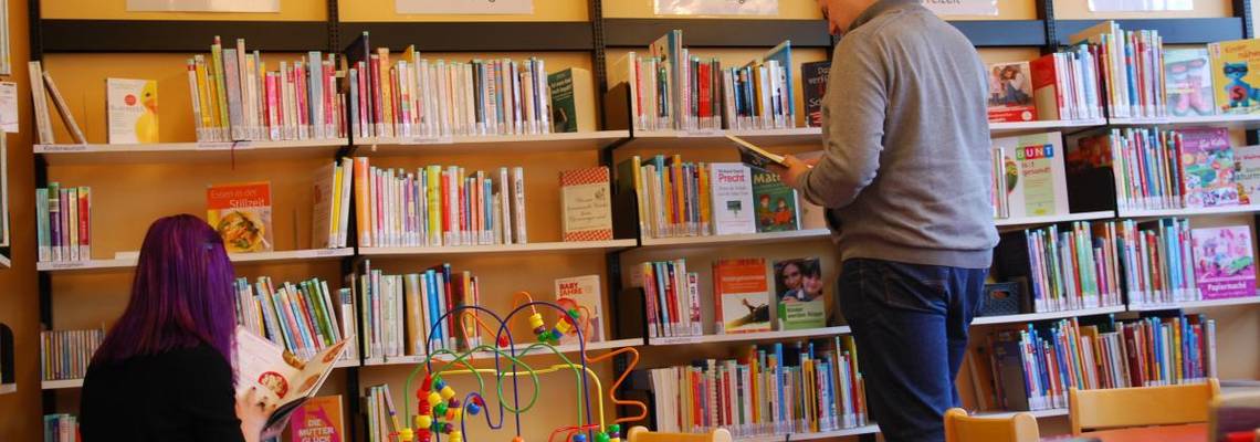 Bücherregale der Elternbibliothek mit zwei Nutzenden vor den Regalen und Kinderbücher im Vordergrund